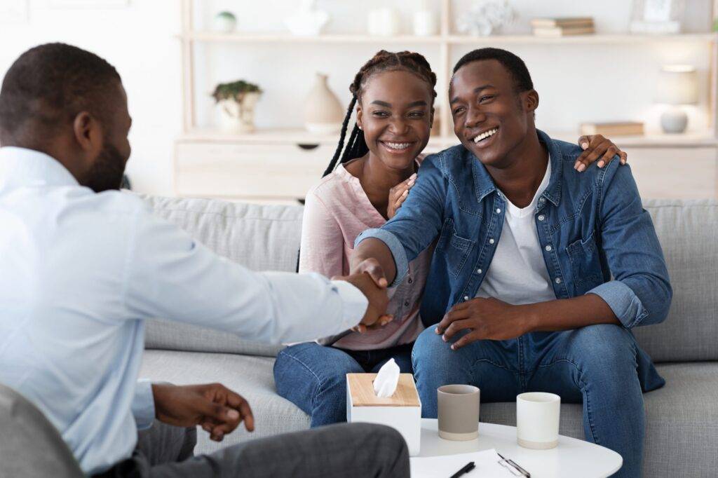 Happy Black Spouses Thankful To Family Counselor After Successful Marital Therapy Session, Shaking Hands With Psychotherapist, Grateful For Reconciliation, Sitting On Couch At Office, Free Space