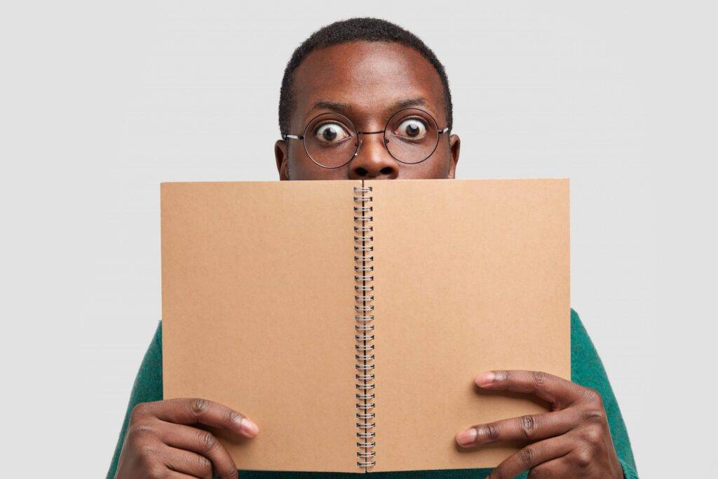Close up shot of shocked black man covers face with spiral notepad, feels astonished, models over white studio background, reads notes written in notebook, wears round glasses, has scared look