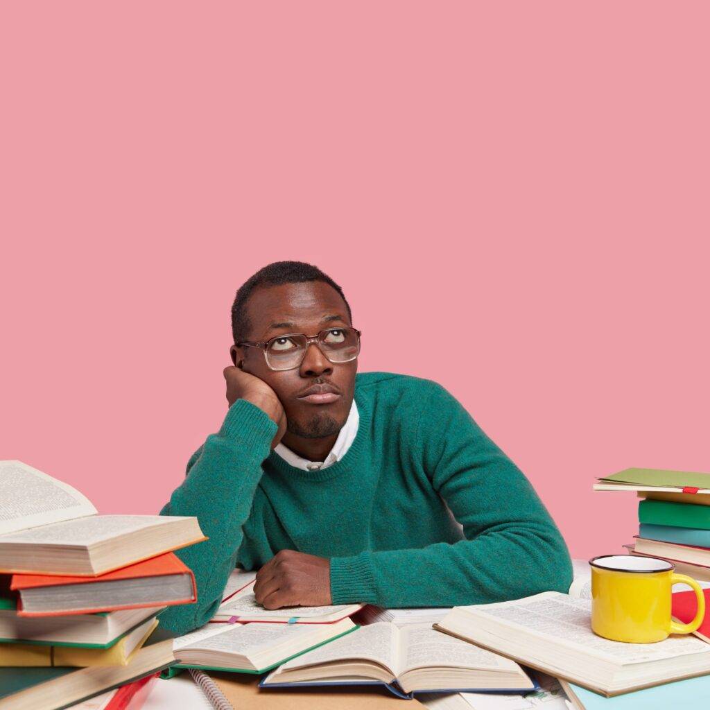 Thoughtful bored black man holds hand on cheek, looks upwards, wears green sweater, optical glasses, thinks about creating new project work, studies indoor with many books, isolated on pink wall