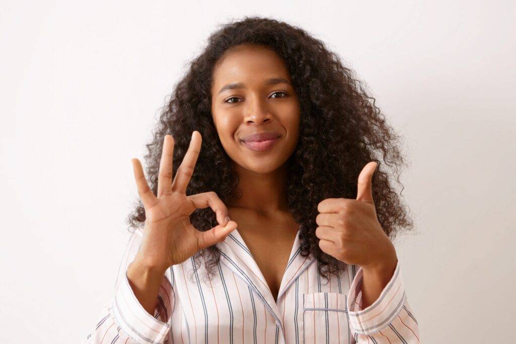 Beautiful happy young mulatto girl with curly black hair posing isolated dressed in silk pajamas, making thumbs up gesture and showing ok sign, smiling.