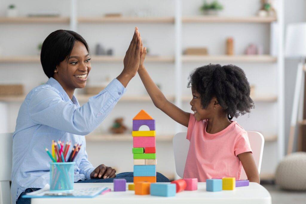Pretty young african american woman preschool teacher exercising with happy curly little girl, sitting at table together, making constructions with colorful wooden play bricks, giving high five