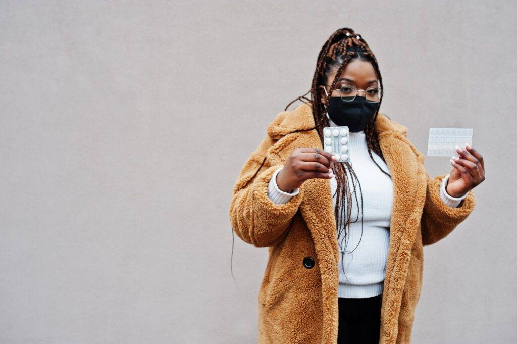 African american woman in eyeglasses and black protective face mask show pills.