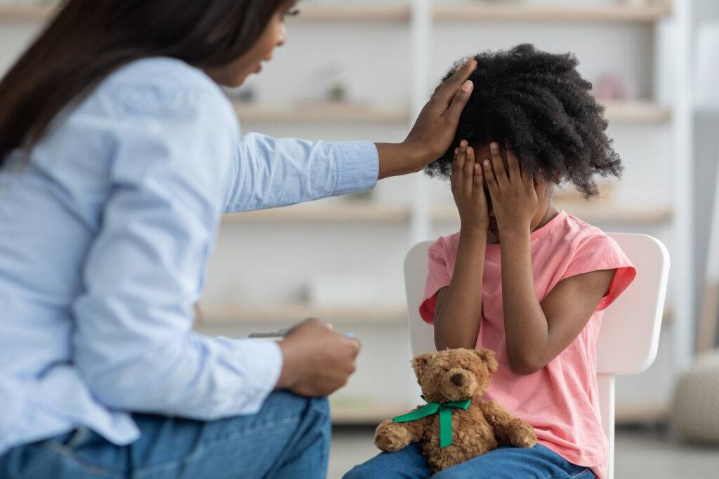 Back view of unrecognizable african american woman comforting emotional curly girl preschooler crying during therapy session with child psychologist, carrying teddy bear on her lap, clinic interior